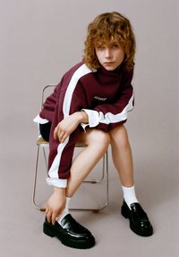 Bordeauxrød sweatshirt med hvide striber, kombineret med skinnende sorte loafers og hvide crew-sokker. Model siddende på en stol med en afslappet positur.