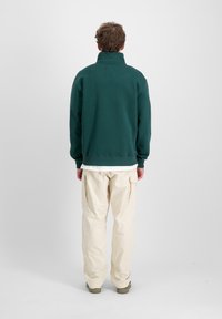 Felpa verde con colletto alto e polsini a coste, abbinata a pantaloni cargo beige chiaro con tasche laterali e vestibilità rilassata.