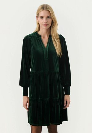 Abito in velluto verde scuro con scollatura a V, maniche lunghe a palloncino e orlo a strati con volant. Superficie liscia e lieve lucentezza in tutto l'abbigliamento.