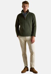 Giovane uomo che indossa un pullover verde oliva a mezze zip, camicia azzurra, pantaloni beige e scarpe eleganti marroni, in piedi con le mani in tasca.