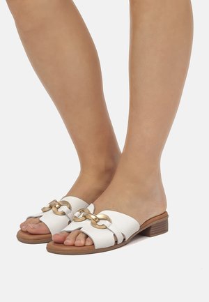 Piedi che indossano sandali bianchi aperti con dettagli dorati a catena e tacchi bassi a blocco in legno su sfondo bianco.