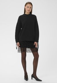 Pull en maille noir à manches longues avec des détails texturés sur les côtés. Associé à une jupe noire à paillettes en couches et des escarpins noirs.