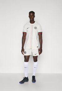 Hvid fodboldtrøje med røde og grønne detaljer, der har Chelsea FC-logoet, parret med hvide shorts og sorte støvler.