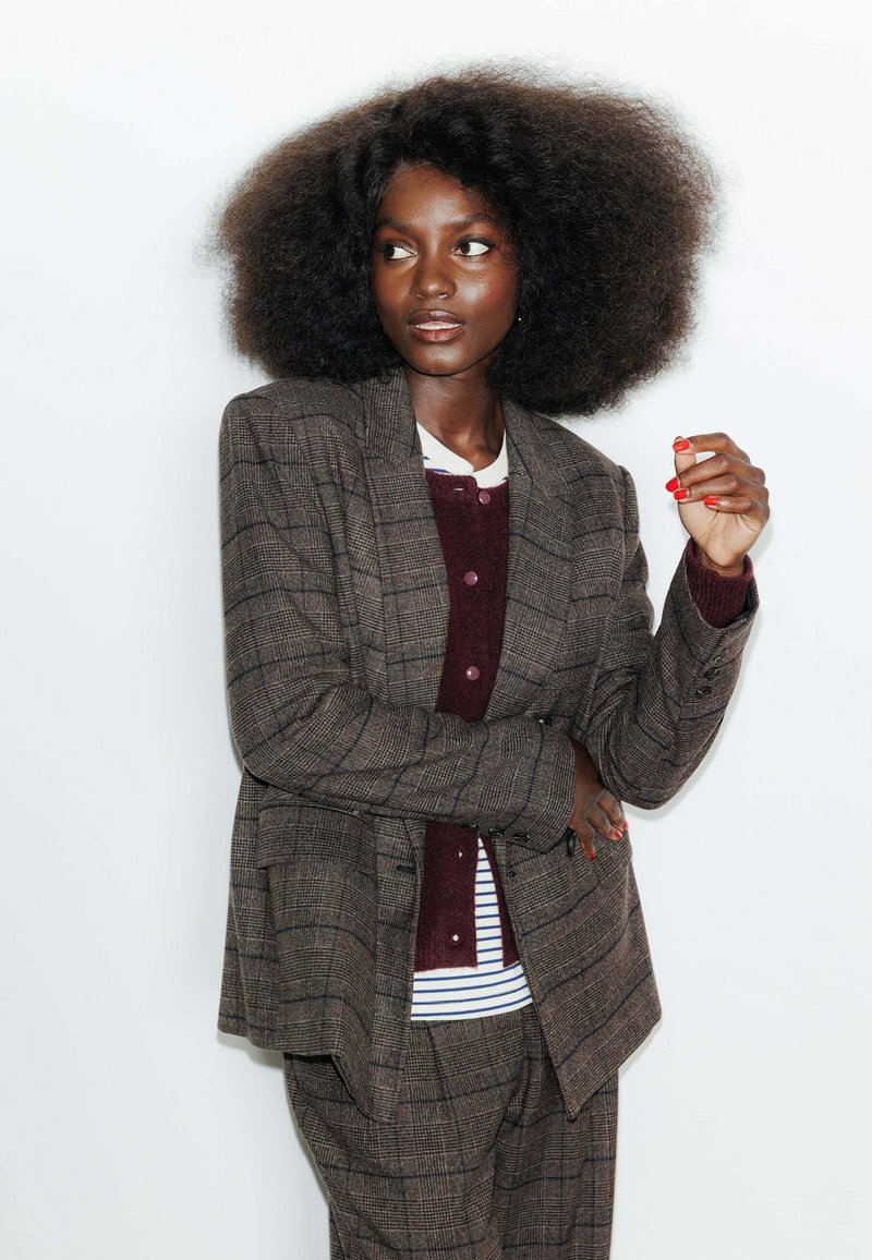 Costume deux pièces à carreaux marron avec un blazer ajusté et un pantalon assorti. Superposé avec une chemise à boutons bordeaux et une chemise rayée en dessous.