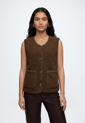 Femme aux longs cheveux foncés porte un gilet marron texturé sans manches, boutonné avec des poches avant, et un pantalon foncé sur un fond uni.
