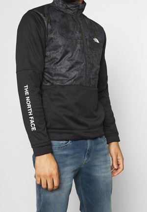 Mężczyzna noszący czarny pulower z suwakiem do ćwiartki, z teksturowanym górnym panelem i napisem "THE NORTH FACE" na rękawie, w niebieskich dżinsach.