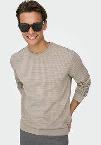 Pull beige texturé avec poignets et col côtelés. Porté avec des lunettes de soleil foncées, dotées d'une monture rectangulaire et de verres noirs.