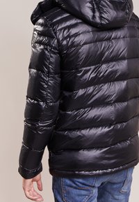 Sort pufferjakke med en skinnende, quiltet overflade, hætte og logo-patch på skulderen. Båret over blå jeans.