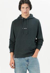 Donkergroene katoenen hoodie met een voorzak, verstelbare capuchon en een wit geborduurd logo op de borst. Model draagt lichtblauwe jeans.