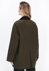 Femeie cu părul blond, îmbrăcată într-o jachetă supradimensionată de culoare verde închis, cu guler negru, văzută din spate pe un fundal alb.
