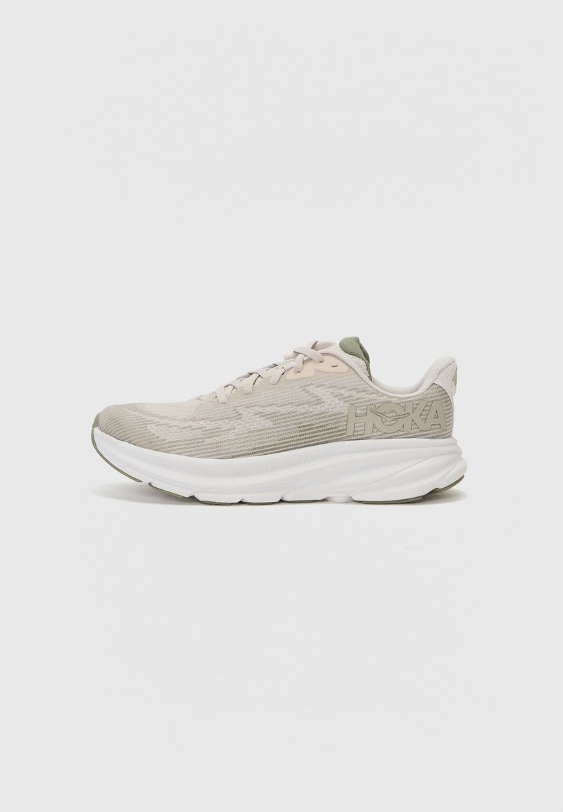 Beige athletic shoe with textured mesh upper, green accents, and thick white sole. Features lace-up design and logo detail on the side.