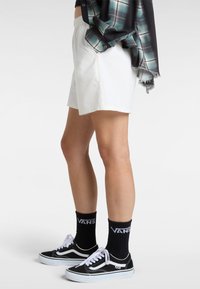 Weiße Shorts mit geometrischem Muster, seitlichen Taschen, kombiniert mit schwarzen Vans Sneakers und schwarzen Crew-Socken mit einem Logo, ergänzt durch ein kariertes Hemd.