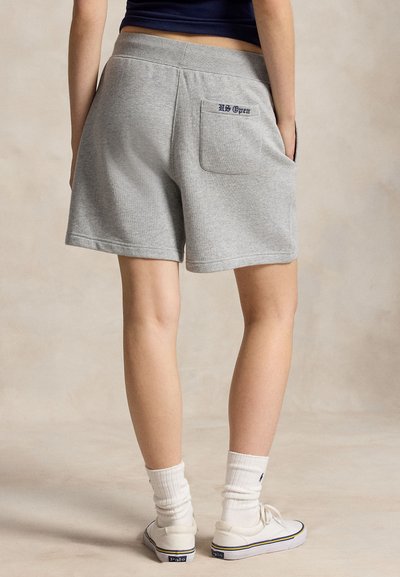 Personne portant un short décontracté gris avec une poche arrière étiquetée "US Open,", des chaussettes blanches montantes, des baskets blanches, debout sur un fond neutre.