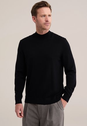 Homme aux cheveux courts châtain clair portant un pull noir à manches longues et un pantalon gris, debout avec une main dans la poche, regardant vers la droite.