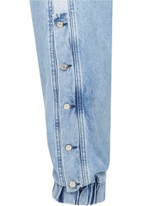 Mâneca unei jachete din denim de culoare albastru deschis, cu manșetă plisată, având nasturi cu clip metalici pe seamă și detalii de cusătură în contrast.