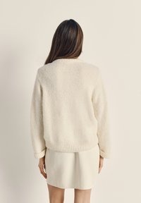 Krémový, texturovaný svetr z vlny s kulatým výstřihem a žebrovaným lemem, zkombinovaný se světle béžovou sukní. Je vidět záda modelky.