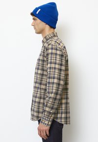 Checkered plaid shirt in beige, brown, and navy with long sleeves. A blue knit beanie is worn. Side profile view against a white background.