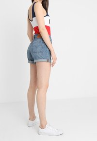 Denim shorts dengan hem yang digulung, menunjukkan warna biru pudar, dipadukan dengan tank top multwarna dan sneakers putih. Patch kulit cokelat di bagian belakang.