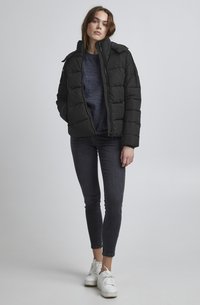 Svart puffjacka med vadderad design, hög krage och dragkedja fram. Bärs över en marinblå tröja, tillsammans med mörka jeans och vita sneakers.