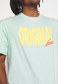 Mintsävyinen puuvillapaita, jossa on rohkeaa keltaista "ORIGINAL"-tekstiä ja punainen "Levi's"-logo. Paita on pyöreällä kaula-aukolla ja lyhyillä hihoilla.