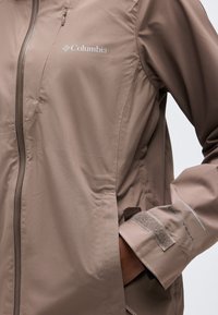 Veste Columbia couleur taupe avec fermeture éclair frontale, poche poitrine, manchette réglable à velcro étiquetée Omni-Tech, et main dans la poche latérale.