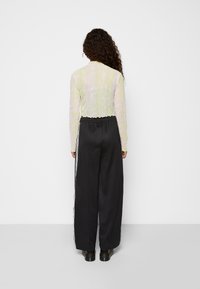 Transparente langärmelige Bluse in hellgelb mit einem Blattmuster; kombiniert mit schwarzen, weit geschnittenen Hosen mit weißen Seitenstreifen.