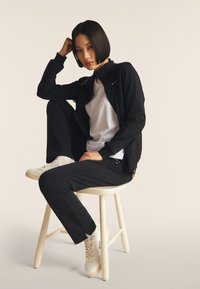 Veste noire zippée et pantalon noir associés à une chemise blanche, assis sur un tabouret blanc. Chaussures en sneakers blanches ; texture lisse.