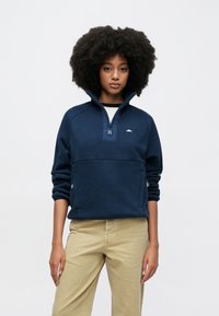 Marineblå fleecegenser med halvlås, glidelåslommer og en diskret logo; kombinert med lyse kaki bukser. Strukturert stoff.