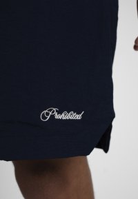 Short bleu marine avec texte blanc en cursive « Prohibited » brodé près de l'ourlet sur la jambe gauche.