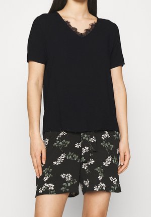 Frau trägt ein schwarzes kurzärmeliges V-Ausschnitt-Oberteil mit Spitzenbesatz und schwarze Shorts mit floralem Muster und gebundenem Taillengürtel.