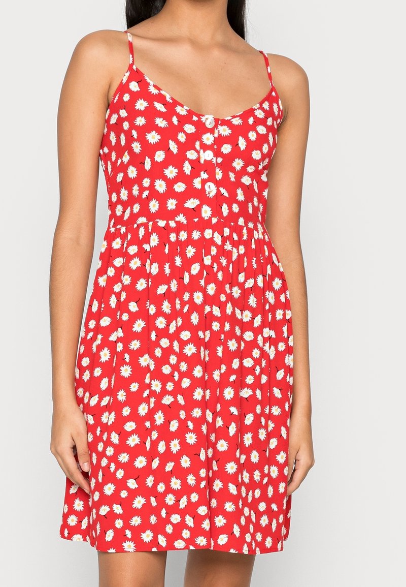 Röd ärmlös klänning med tunna axelband, vit prästkrage-blommönster, framknappar och samlad midja, bärs av en stående kvinna.