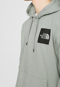 Sudadera gris con cordones ajustables, que presenta un parche de logo cuadrado negro en el lado izquierdo del pecho y un bolsillo tipo canguro en la parte frontal.