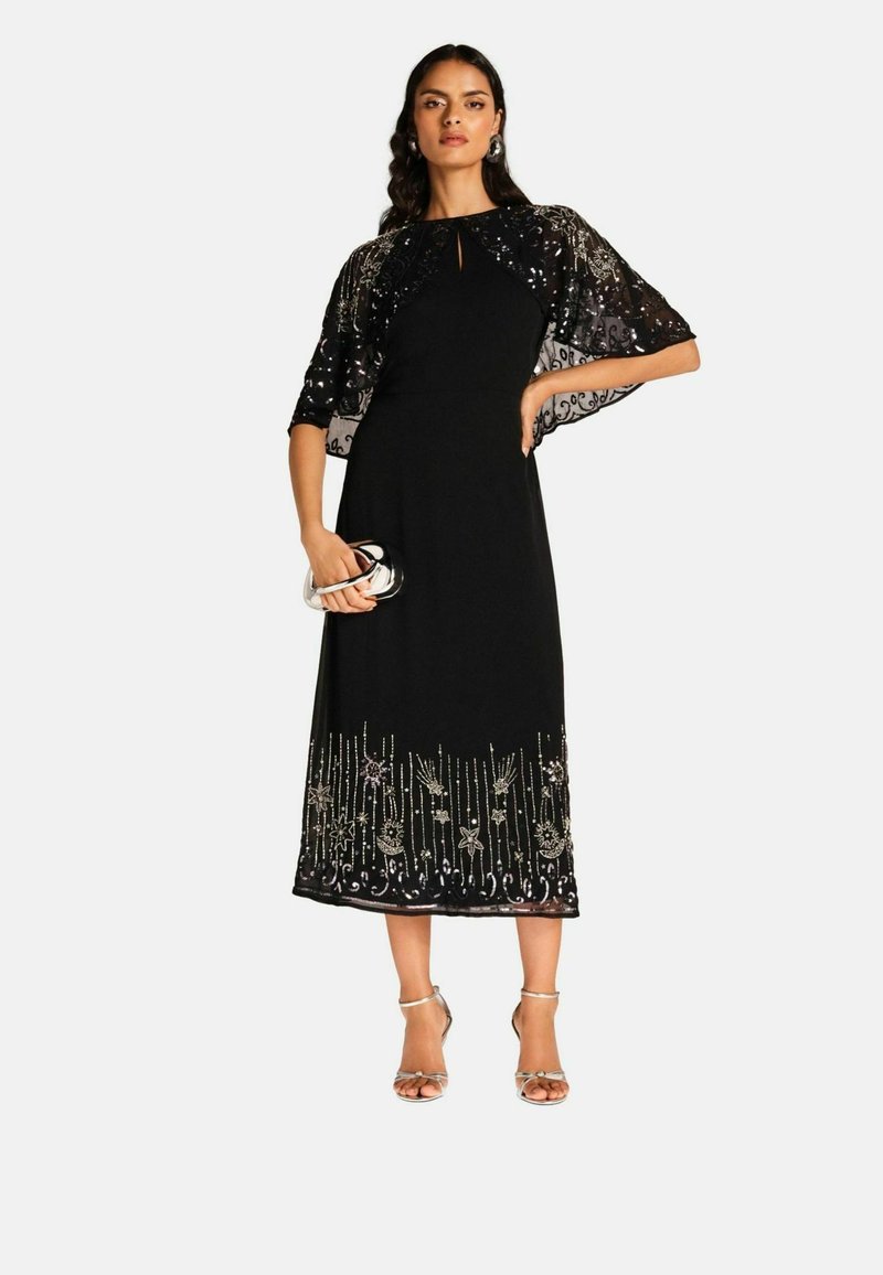 Ilga juoda suknelė su permatomu, karoliukais dekoruotu apdupimu. Išilginiai floristiniai raštai ant apačios. Lygus audinys su prigludusia liemenėle ir laisvai krentančia sijonu.