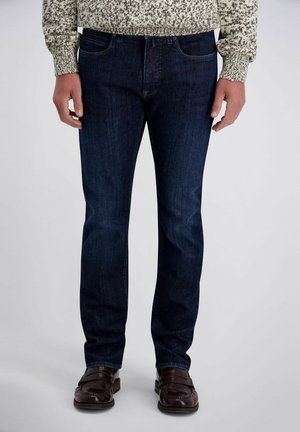 Mann trägt dunkelblaue Jeans, braune Ledermokassins und einen beigen Pullover mit Muster, steht entspannt mit den Händen an den Seiten.