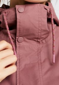 Personne ajustant la capuche de la veste rose avec des cordons tressés et des boutons-pression mats près du cou.