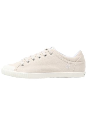 Nízký béžový plátěný teniska s bílou gumovou podrážkou, kovovými dírkami na tkaničky, šněrováním vpředu a nenápadným vyšitým logem na boku.