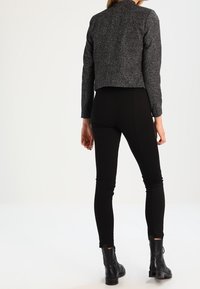 Femme portant une veste grise courte, un pantalon noir ajusté et des bottines noires, debout de dos face à l'appareil photo.