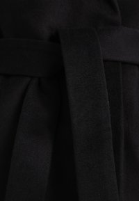 Schwarzer Wollstoff mit glatter Textur, ausgestattet mit einem gebundenen Gürtel. Das Design umfasst sanfte Falten, die die strukturierte Form betonen.