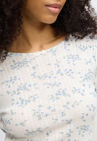 Haut en maille à motif floral avec une base crème ornée de fleurs bleues et vertes, texture côtelée, et un petit nœud décoratif au niveau de l'encolure.