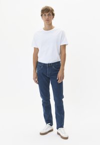 Modri jeans iz denima s petimi žepi, klasičnega kroja in s gumbom, v kombinaciji z enostavno belo majico in belimi športnimi copati.
