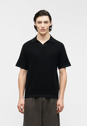 Jeune homme aux cheveux courts et foncés, portant une chemise noire à manches courtes avec col et un pantalon marron ample, debout devant un fond blanc uni.