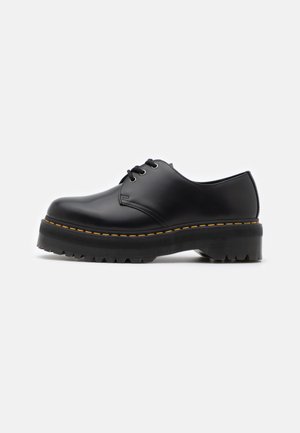 Chaussure noire en cuir à lacets avec un bout arrondi, semelle en caoutchouc noir épais et surpiqûres jaunes le long du bord. Présente une texture lisse et un design solide.