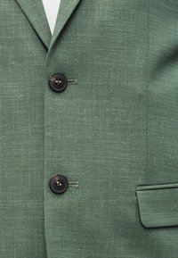 Blazer vert texturé avec deux boutons noirs fermés, un revers cranté et une poche à rabat du côté droit.