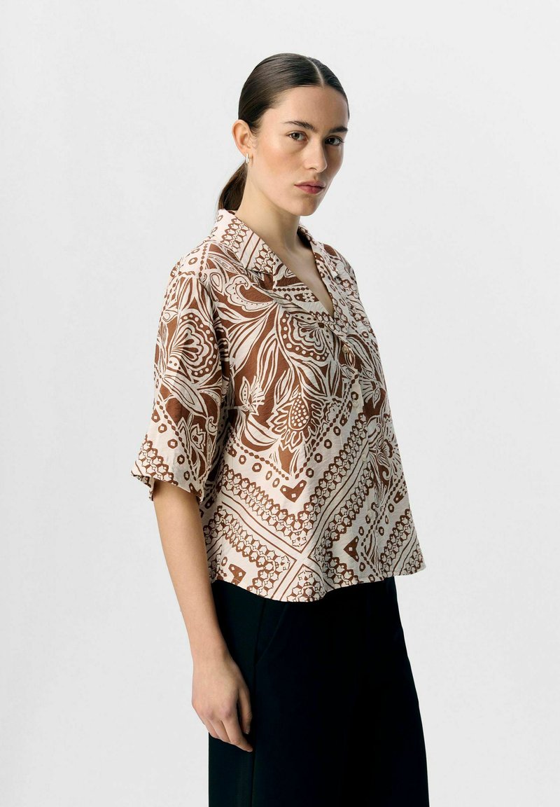 Camicia a maniche corte marrone con stampa paisley, dotata di colletto e una vestibilità loose, realizzata in tessuto leggero, abbinata a pantaloni neri.