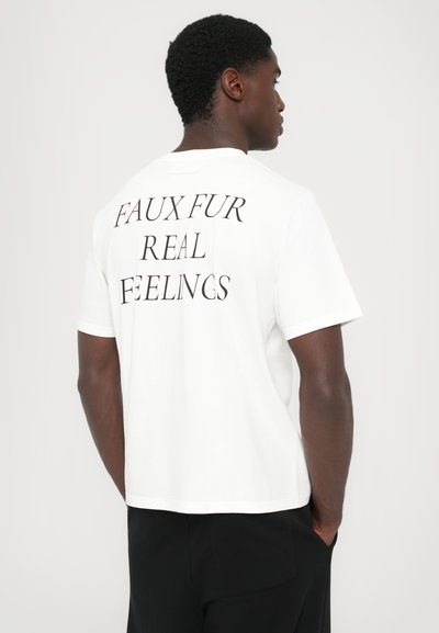 T-shirt de algodão branco com o texto "FELTROS FALSOS, SENTIMENTOS REAIS" em fonte preta em negrito nas costas. Mangas curtas e um ajuste descontraído.