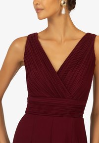 Burgunderfarbenes, strukturiertes Kleid mit gepflüschtem Design, V-Ausschnitt und taillierter Passform. Es hat ein glattes Band an der Taille und elegante Ohrringe.