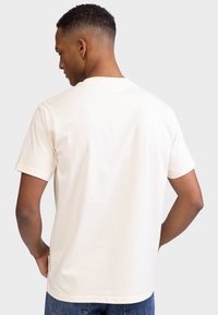T-shirt bege de algodão com mangas curtas, gola redonda e um pequeno detalhe de etiqueta na lateral. O tecido tem uma textura suave.
