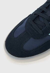 Sapatilha azul-marinha com materiais de camurça e nylon, apresentando uma textura suave, biqueira redonda e um destaque em azul claro contrastante na lateral.