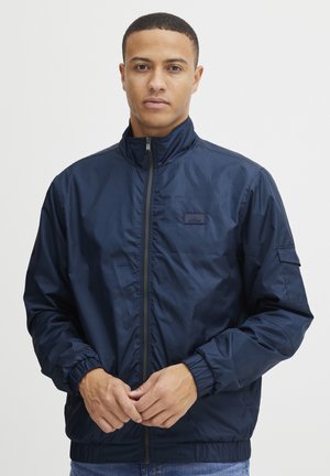 Chaqueta ligera de color azul marino con cuello alzado, cremallera frontal, puños elásticos y un detalle de etiqueta en el pecho. Material suave y resistente al agua.