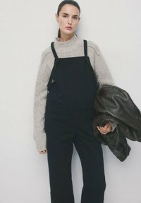 Schwarze Jeans-Latzhose mit verstellbaren Trägern, kombiniert mit einem hellgrauen Strickpullover. Das Modell hält eine dunkle Lederjacke. Minimalistisches Design.